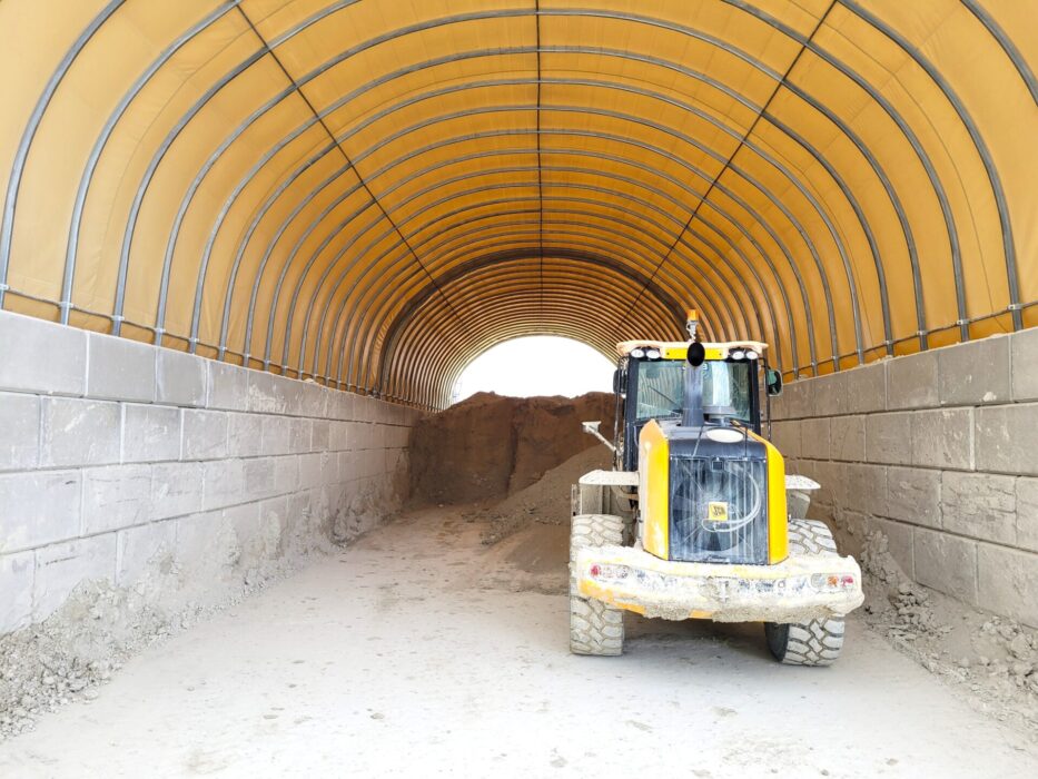 W hali łukowej do składowania kredy widoczny jest żółty ładowacz czołowy stojący na utwardzonym podłożu. Ściany z betonowych bloków oraz żółte poszycie namiotowe zapewniają ochronę i stabilność konstrukcji. W głębi hali znajduje się pryzma materiału.