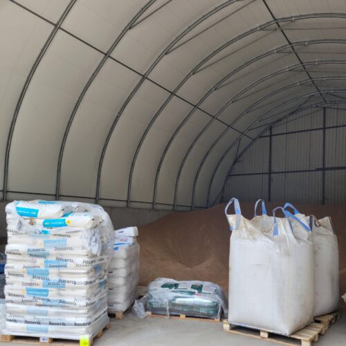 Na zdjęciu znajduje się hala namiotowa z łukową konstrukcją stalową i jasnym poszyciem. Wewnątrz widać składowane zboże usypane w pryzmy przy ścianie z blachy. Z przodu ustawione są palety z workami i big-bagami – część z nich to pasze lub dodatki paszowe zapakowane w oryginalne opakowania. Całość wskazuje na wykorzystanie hali zarówno jako magazynu zboża, jak i miejsca do przechowywania pasz.