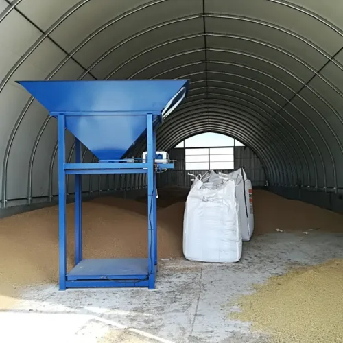 Wnętrze hali magazynowej z niebieskim systemem załadunkowym i przechowywanymi workami big-bag. Konstrukcja stalowa tworzy tunel magazynowy, a na podłodze widoczne są resztki ziarna po procesach magazynowych.