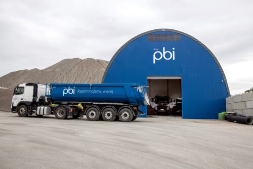 Hala garażowa w kolorze niebieskim, wykorzystywana do przechowywania ciężarówek i pojazdów transportowych. Przed halą stoi samochód ciężarowy z naczepą w barwach firmowych PBI.