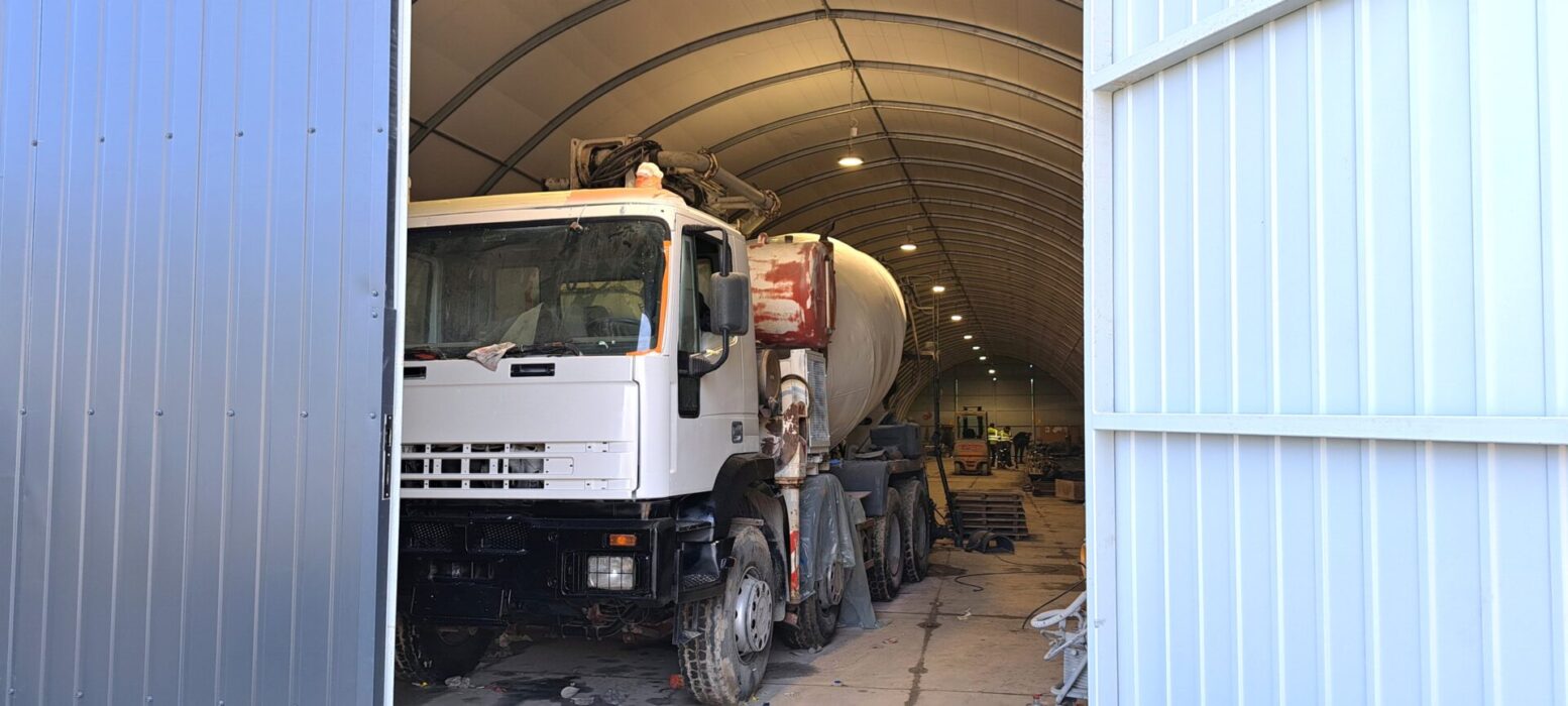 Wnętrze dużej hali namiotowej z betonową posadzką, w której stoi betoniarka i inne maszyny budowlane, oświetlone równym rzędem lamp sufitowych.