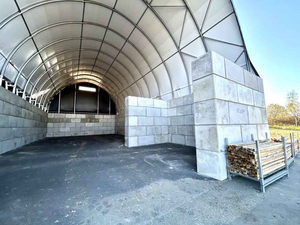 Nowoczesna hala łukowa z betonowymi ścianami wewnętrznymi, przeznaczona do składowania grysu i kruszywa. Konstrukcja z plandeki na stalowym szkielecie zapewnia trwałą ochronę materiałów przed warunkami atmosferycznymi. Obiekt przygotowany do użytkowania w branży budowlanej i drogowej.
