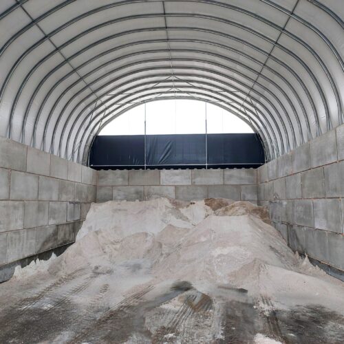 Wewnątrz hali łukowej znajdują się duże pryzmy soli drogowej i piasku. Konstrukcja opiera się na wysokich ścianach z betonowych bloków oraz stalowym szkielecie pokrytym jasnym poszyciem namiotowym. Na podłożu widoczne są ślady po pracy maszyn transportujących materiał.