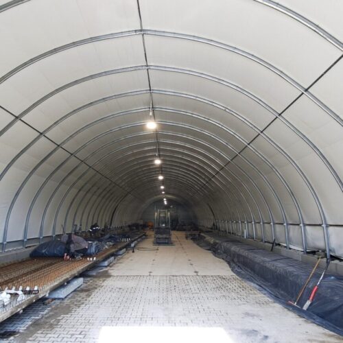 Wnętrze hali namiotowej o konstrukcji łukowej, wykorzystywanej jako przestrzeń produkcyjna z rozłożonymi elementami zbrojenia i materiałami budowlanymi.