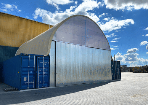 Hala namiotowa zamontowana jako zadaszenie nad dwoma kontenerami morskimi, tworząca funkcjonalną przestrzeń magazynową. Konstrukcja opiera się na metalowym stelażu i półprzezroczystym poszyciu, a całość uzupełniają przesuwne drzwi z blachy trapezowej. Obiekt znajduje się na utwardzonym placu przemysłowym.