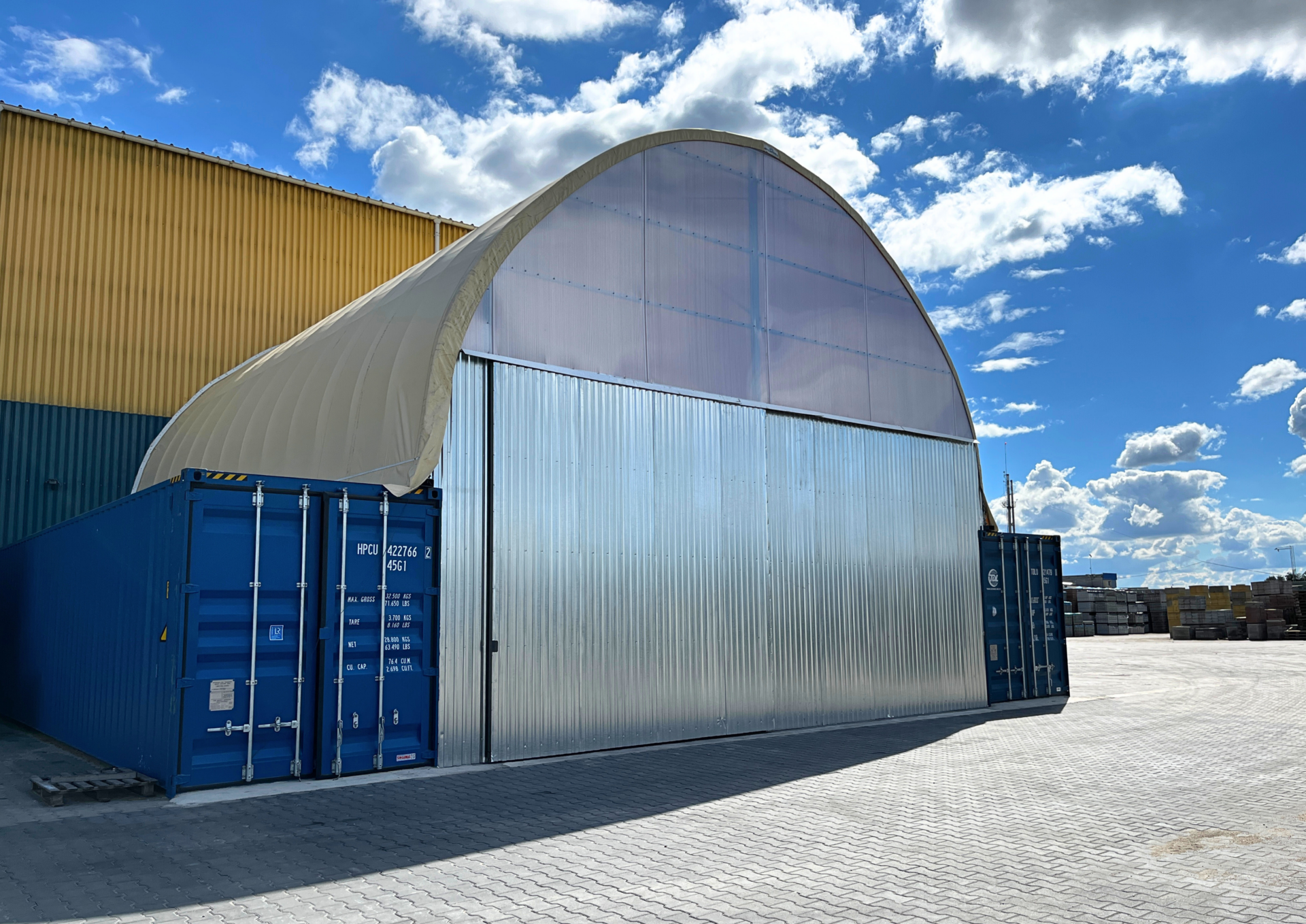 Hala namiotowa zamontowana jako zadaszenie nad dwoma kontenerami morskimi, tworząca funkcjonalną przestrzeń magazynową. Konstrukcja opiera się na metalowym stelażu i półprzezroczystym poszyciu, a całość uzupełniają przesuwne drzwi z blachy trapezowej. Obiekt znajduje się na utwardzonym placu przemysłowym.
