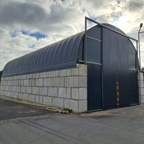 Na zdjęciu widoczna jest hala łukowa wypełniona dużymi pryzmami soli drogowej i piasku. Konstrukcja opiera się na stalowym szkielecie pokrytym jasnym poszyciem namiotowym, a wnętrze hali chroni materiał przed deszczem i wiatrem. Przed obiektem widać utwardzoną nawierzchnię z odciśniętymi śladami maszyn.