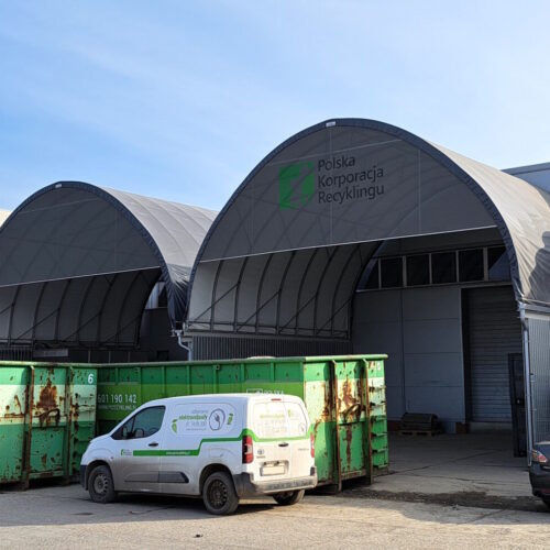 Na zdjęciu widoczne są łukowe hale magazynowe z tworzywa typu Shelterall z logo „Polska Korporacja Recyklingu” na froncie. Przed obiektem znajdują się zielone kontenery, z których jeden wypełniony jest odpadami wielkogabarytowymi, a na placu zaparkowane są dwa pojazdy, w tym biały samochód firmowy z oznaczeniami dotyczącymi odbioru elektroodpadów. Obok hal znajduje się również podest z instalacją przemysłową.