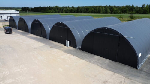 Zespół dużych hal łukowych ustawionych w rzędzie, każda z ciemnym poszyciem i zamkniętymi bramami. Konstrukcje stoją na utwardzonym placu, a w tle widać tereny zielone i zabudowania gospodarcze.