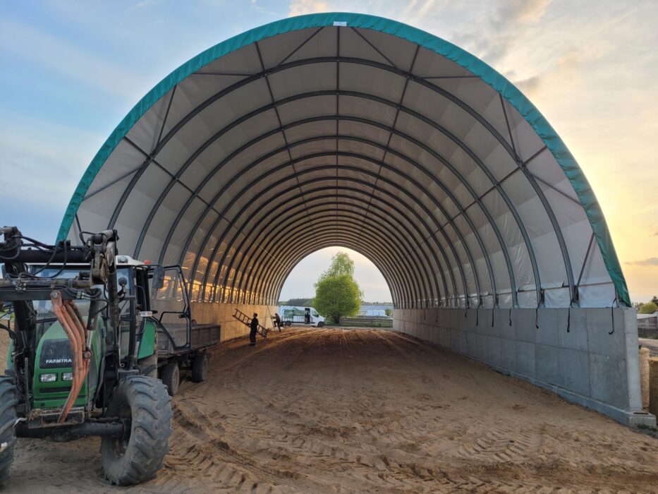 Hala tunelowa o konstrukcji łukowej, oparta na betonowych ścianach oporowych. Dach wykonany z jasnej membrany PCV z zielonym pasem na krawędzi. Wewnątrz znajduje się piaszczyste podłoże, a przy wjeździe ustawiony jest ciągnik rolniczy z przyczepą.