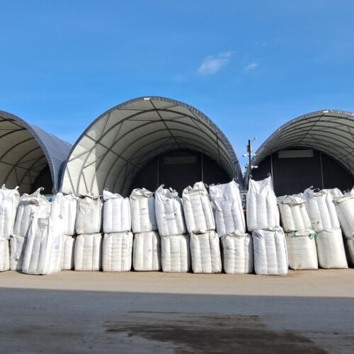 Na zdjęciu widoczne są hale łukowe typu Shelterall, służące jako zadaszone magazyny dla odpadów lub materiałów sypkich. Przed nimi ustawione są rzędy białych worków typu big bag, częściowo wypełnionych. Całość zlokalizowana jest na utwardzonym placu przemysłowym, co wskazuje na zorganizowaną przestrzeń do składowania i logistyki.