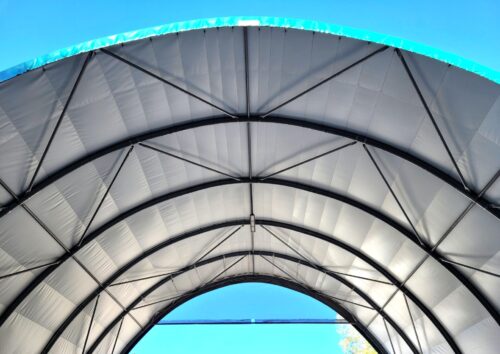 Na zdjęciu widoczna jest konstrukcja dachu hali łukowej z membrany PCV rozpiętej na stalowych ramach. Wyraźnie zaznaczone są łuki nośne oraz krzyżowe wzmocnienia, które stabilizują całą konstrukcję. Jasne poszycie przepuszcza naturalne światło, zapewniając dobre doświetlenie wnętrza, a jednocześnie chroni przed czynnikami atmosferycznymi.