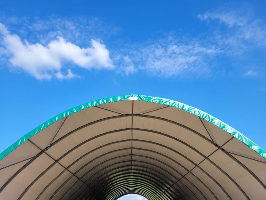 Na zdjęciu widzimy halę tunelową o konstrukcji łukowej, wykonaną z ocynkowanych profili stalowych i pokrytą membraną PCV w jasnym kolorze. Krawędzie poszycia wykończone są zielonym pasem zabezpieczającym, nadającym obiektowi estetyczny wygląd. Konstrukcja wyróżnia się prostą, aerodynamiczną formą, która zapewnia trwałość i odporność na warunki atmosferyczne.