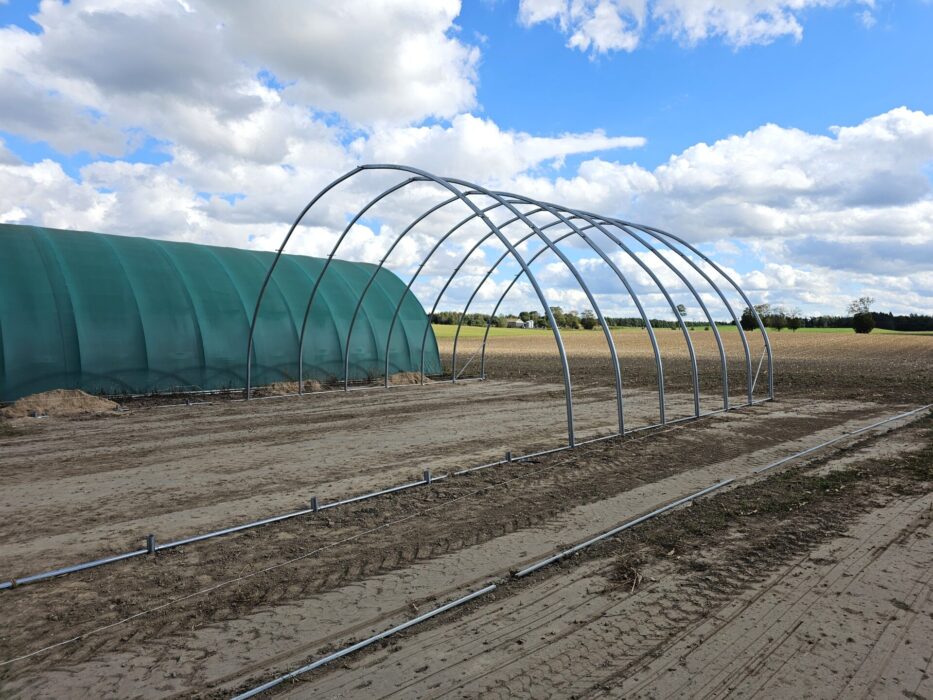 Stalowa konstrukcja łukowa w trakcie montażu, przygotowana pod zadaszenie membranowe. Obok stoi gotowa hala tunelowa pokryta zieloną plandeką.