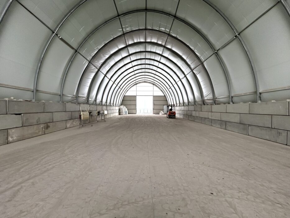 ChatGPT powiedział:  Wnętrze hali łukowej z betonowymi blokami ustawionymi po bokach jako ściany oporowe. Na podłożu znajduje się warstwa drobnego materiału sypkiego, a w głębi ustawiony jest wózek widłowy.