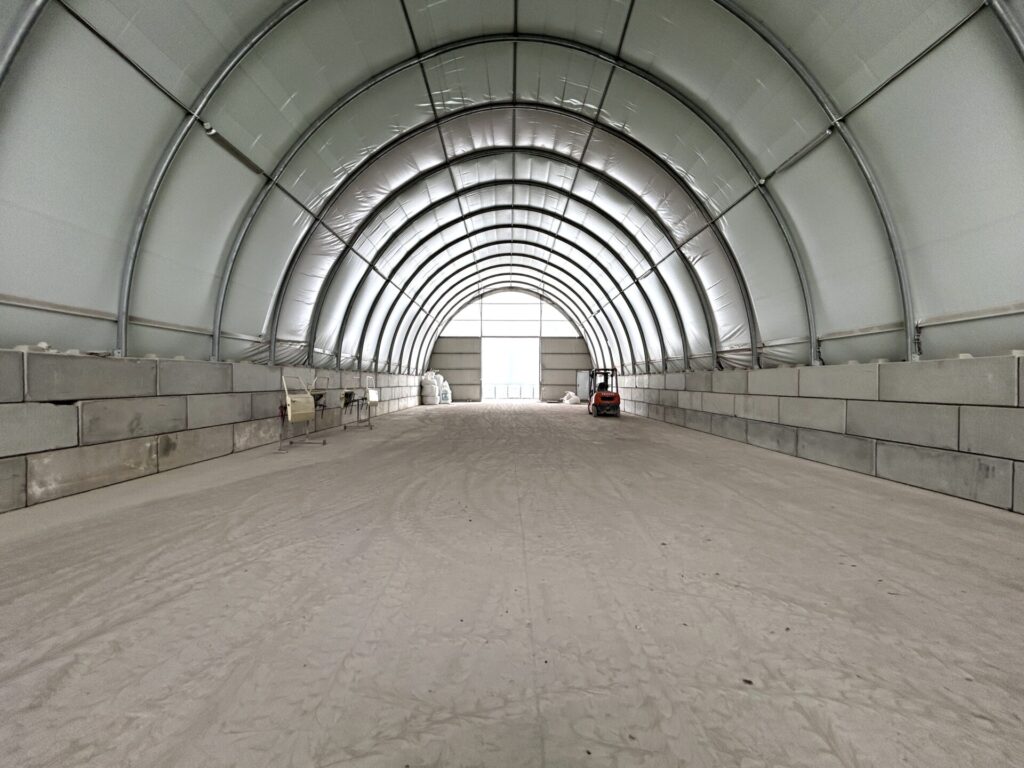 Wnętrze hali łukowej z betonowymi blokami ustawionymi po bokach jako ściany oporowe. Na podłożu znajduje się warstwa drobnego materiału sypkiego, a w głębi ustawiony jest wózek widłowy.