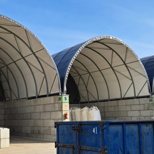 Na zdjęciu widoczne są łukowe hale magazynowe z betonowymi bokami, oznaczone numerami, wykorzystywane do przechowywania materiałów w big bagach. Przed halami ustawione są liczne białe worki oraz niebieski kontener. Całość znajduje się na utwardzonym placu przemysłowym, w tle widoczne są silosy przemysłowe.