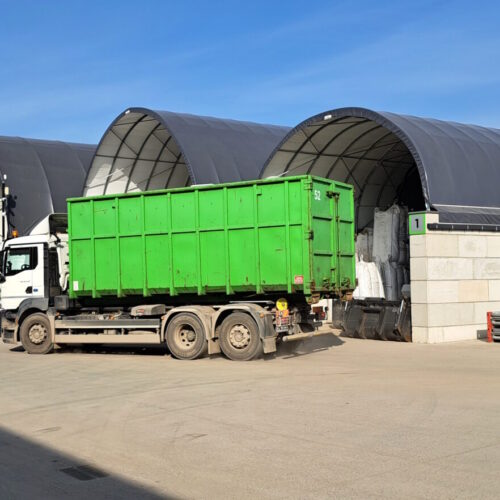 Na zdjęciu widoczne są łukowe hale magazynowe typu Shelterall, ustawione w rzędzie na utwardzonym placu przemysłowym. Przed jedną z hal stoi ciężarówka z zielonym kontenerem, a obok niej znajdują się duże worki typu big bag oraz elementy stalowe. Całość otoczona jest betonowym ogrodzeniem, co wskazuje na zorganizowaną przestrzeń składowo-logistyczną.