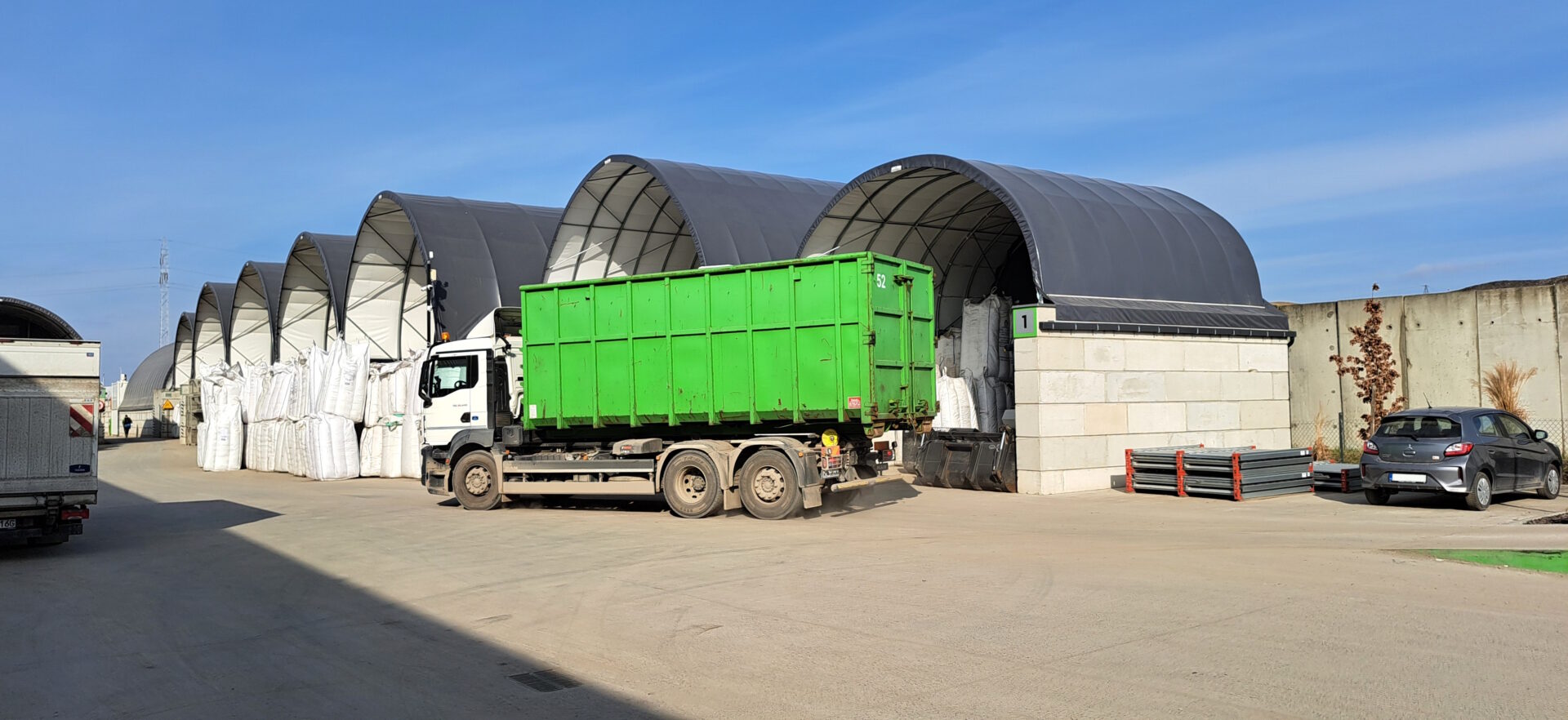Na zdjęciu widoczne są łukowe hale magazynowe typu Shelterall, ustawione w rzędzie na utwardzonym placu przemysłowym. Przed jedną z hal stoi ciężarówka z zielonym kontenerem, a obok niej znajdują się duże worki typu big bag oraz elementy stalowe. Całość otoczona jest betonowym ogrodzeniem, co wskazuje na zorganizowaną przestrzeń składowo-logistyczną.