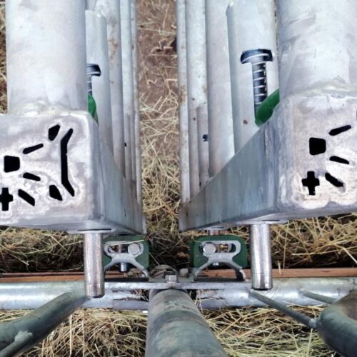 Zbliżenie na metalowe elementy ogrodzenia z regulowanymi zamkami i sprężynami. W tle widać siano rozłożone na ziemi. Konstrukcja wygląda na część systemu do zabezpieczania lub regulacji przegród w budynku inwentarskim.