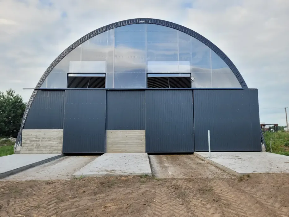 Hala łukowa 15x30 metrów z nowoczesnym wykończeniem i dużymi bramami wjazdowymi, osadzona na betonowej podmurówce. Konstrukcja jest przystosowana do intensywnego użytkowania, zapewniając trwałość i ochronę dla przechowywanych materiałów lub sprzętu.