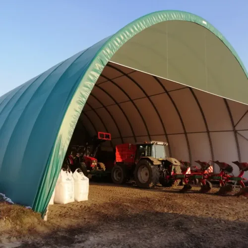 Otwarta hala łukowa 15x30 pełniąca wiatę na maszyny rolnicze. W środku stoi traktor z pługiem oraz worki big-bag przy wjeździe.