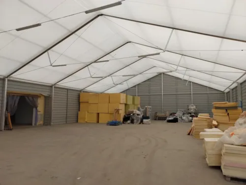 Wnętrze hali dwuspadowej. Pod sklepieniem widać stelaż, który pozwala na rozprowadzenie materiału PVC jako pokrycia dachu. Wewnątrz znajdują się materiały budowlane.