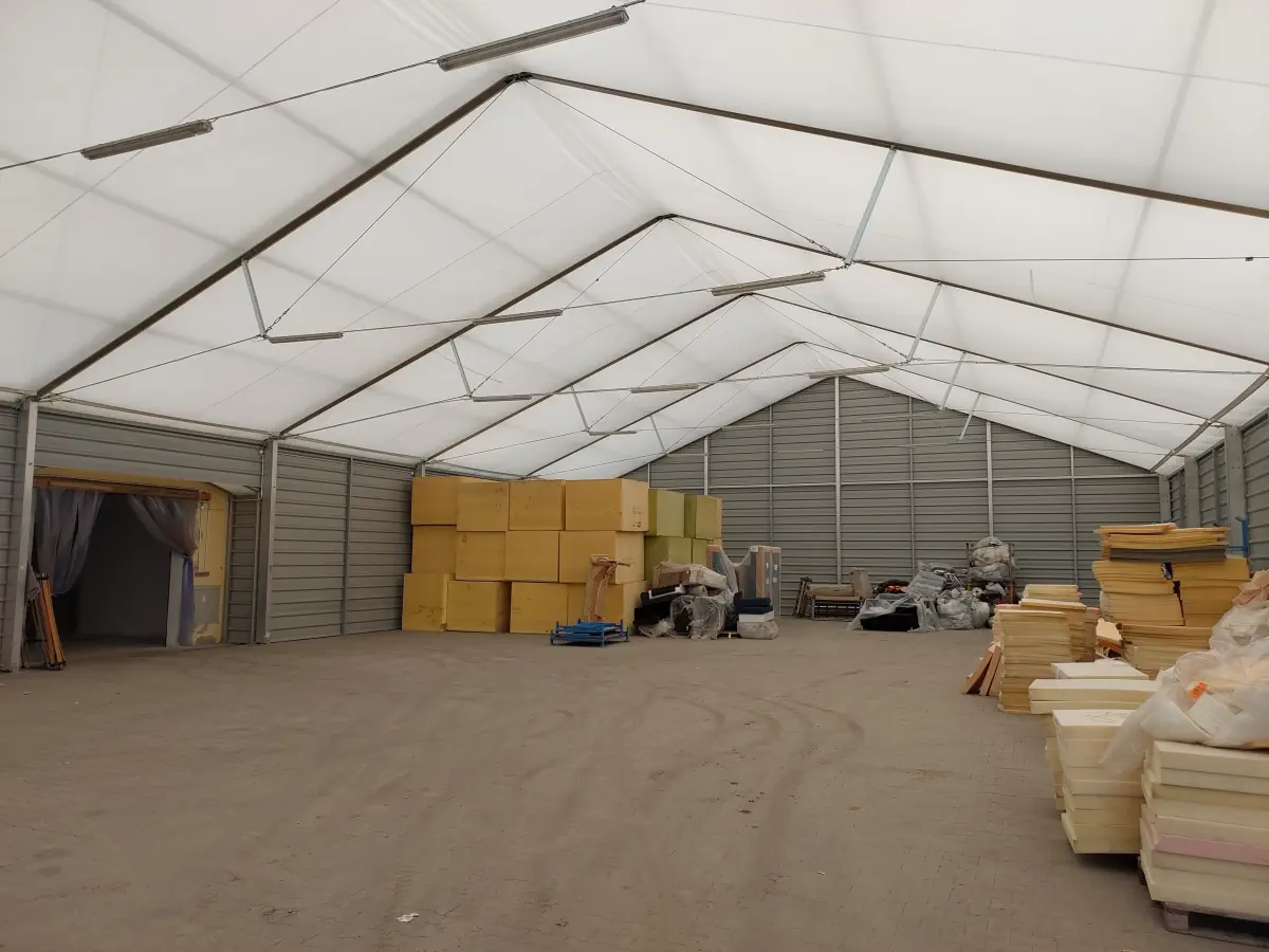 Wnętrze hali dwuspadowej. Pod sklepieniem widać stelaż, który pozwala na rozprowadzenie materiału PVC jako pokrycia dachu. Wewnątrz znajdują się materiały budowlane.
