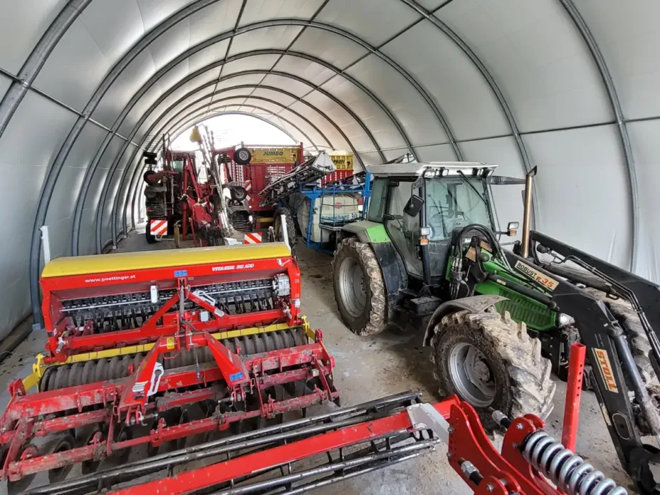 Hala namiotowa 12x30 metrów o wytrzymałej konstrukcji łukowej wykorzystywana jako schronienie dla sprzętu rolniczego. W jej wnętrzu ustawiono ciągniki i różnorodne maszyny, co pokazuje dużą pojemność obiektu.