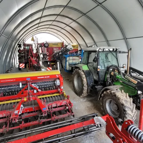 Hala namiotowa 12x30 metrów o wytrzymałej konstrukcji łukowej wykorzystywana jako schronienie dla sprzętu rolniczego. W jej wnętrzu ustawiono ciągniki i różnorodne maszyny, co pokazuje dużą pojemność obiektu.