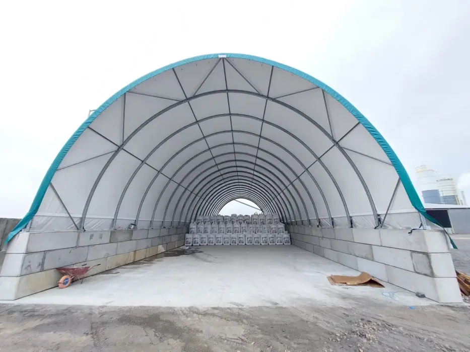 Hala łukowa 15x30 metrów w jasnym kolorze z wysokim wnętrzem, idealna do zastosowań magazynowych i przemysłowych. W środku widoczne są rzędy materiałów w workach, co pokazuje funkcjonalność obiektu.