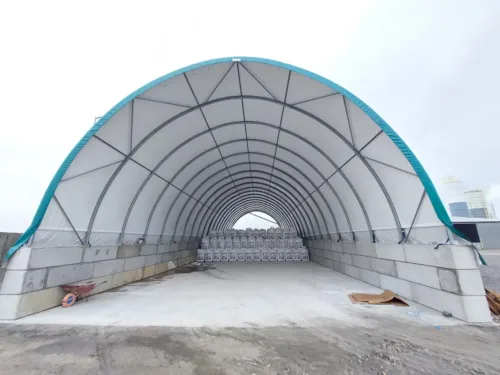 Hala łukowa 15x30 metrów w jasnym kolorze z wysokim wnętrzem, idealna do zastosowań magazynowych i przemysłowych. W środku widoczne są rzędy materiałów w workach, co pokazuje funkcjonalność obiektu.