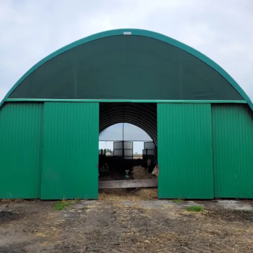 Hala namiotowa 12x30 metrów wyposażona w przesuwne bramy wjazdowe z blachy trapezowej w kolorze zielonym. Konstrukcja pełni rolę magazynu gospodarczego, chroniąc przechowywane wewnątrz zasoby przed deszczem i wiatrem.