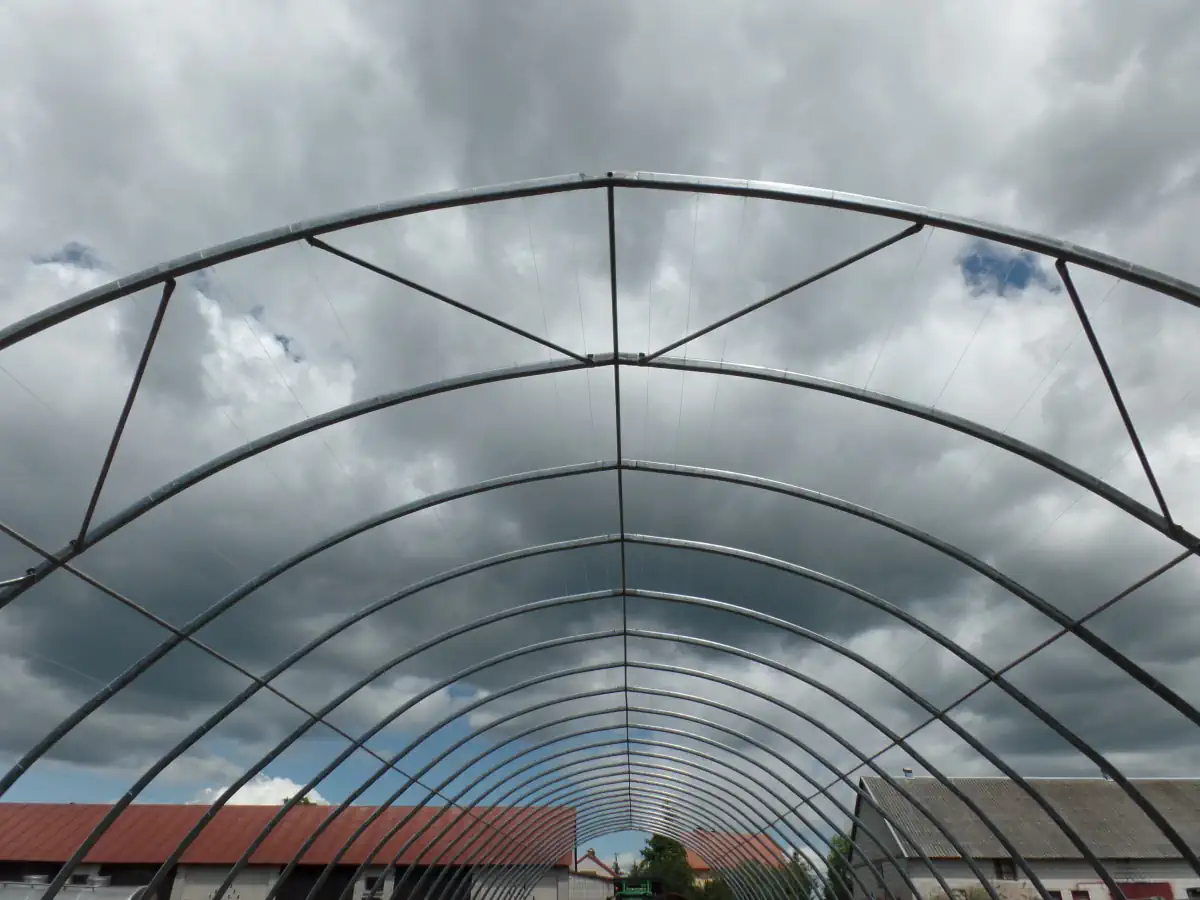 Widok z wnętrza hali namiotowej w trakcie montażu, przedstawiający stalową konstrukcję o łukowym kształcie. Na niebie dominują gęste chmury, a w tle widoczne są zabudowania gospodarstwa. Konstrukcja jest otwarta, bez poszycia plandekowego.