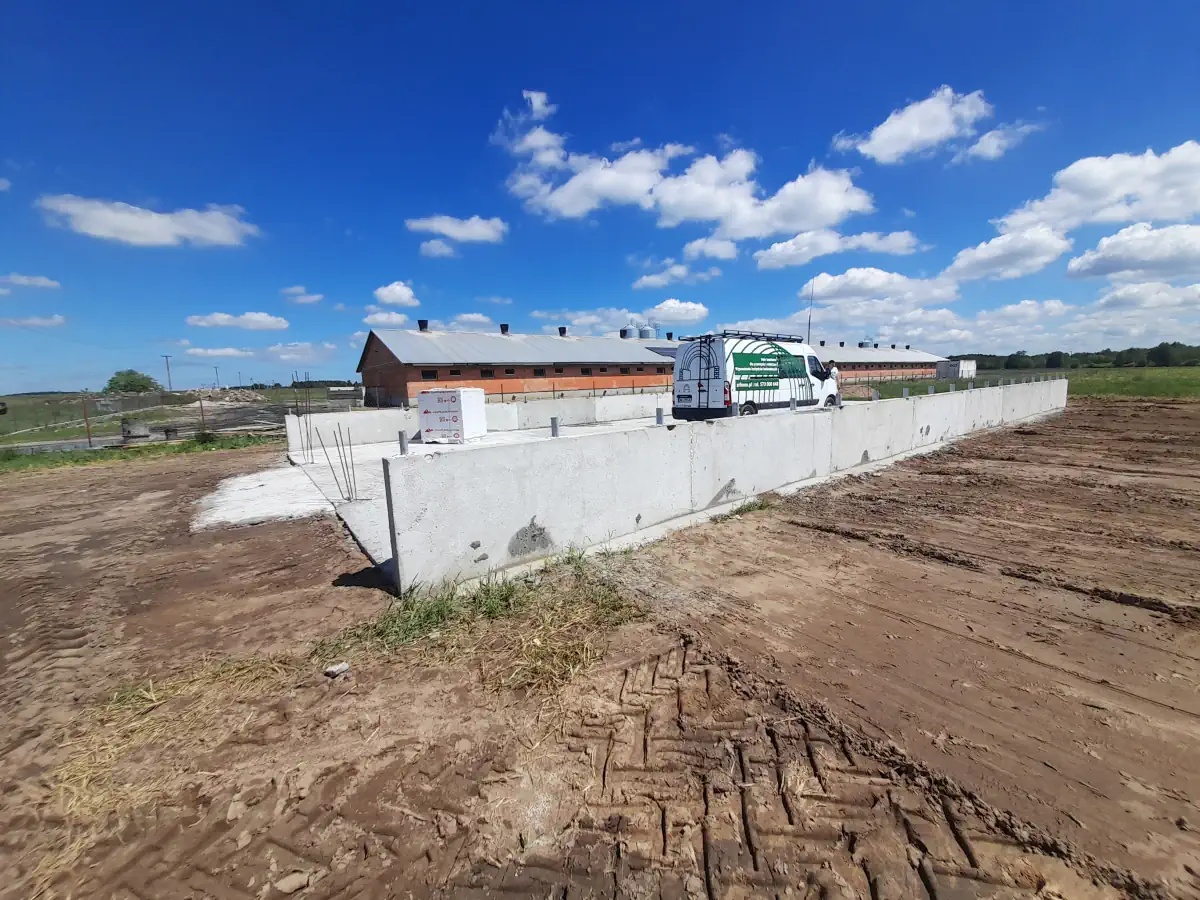Betonowa podstawa przygotowana pod montaż hali namiotowej, ustawiona na wyrównanym terenie. W tle stoi biały samochód dostawczy z grafiką firmy budowlanej oraz długie budynki gospodarcze.