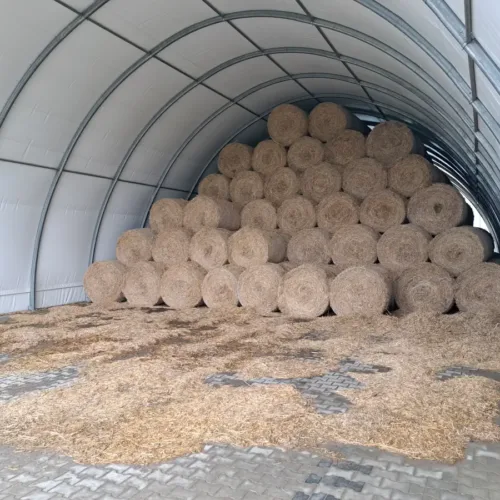 Przestronna hala namiotowa 12x30 metrów z solidną stalową konstrukcją, w której składowane są liczne baloty słomy ułożone w wysokie stosy. Utwardzona posadzka z kostki brukowej ułatwia utrzymanie porządku i wygodne magazynowanie pasz rolniczych.