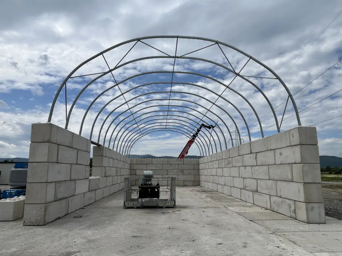 Szkielet stalowy hali namiotowej ustawiony na wysokich ścianach z bloków betonowych. Na podłożu widoczna jest płyta betonowa, a w tle maszyna budowlana.