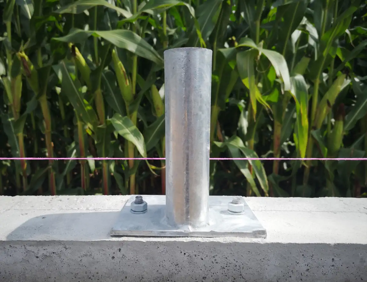 Metalowa stopka montażowa przykręcona do betonowego podłoża, stanowiąca element mocowania konstrukcji. W tle widoczne jest pole kukurydzy i napięta różowa linka wyznaczająca linię montażu.