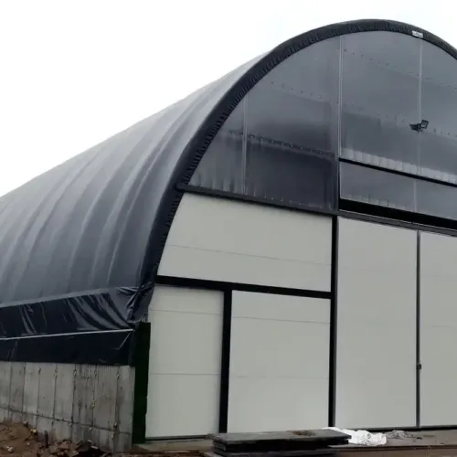 Zabudowana hala namiotowa 12x30 metrów z czarnym poszyciem i dużymi bramami wjazdowymi z paneli, osadzona na betonowej podmurówce. Obiekt pełni funkcję trwałego magazynu gospodarczego, chroniącego sprzęt lub plony przed zmiennymi warunkami pogodowymi.