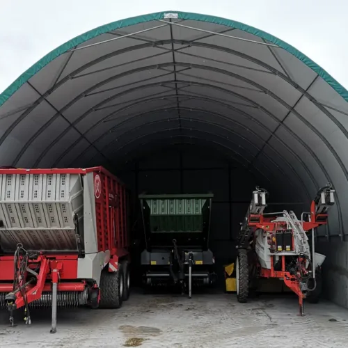Wnętrze hali namiotowej 10x15 z betonowymi ścianami oporowymi i zaparkowanymi maszynami rolniczymi. Konstrukcja łukowa chroni sprzęt przed warunkami pogodowymi.