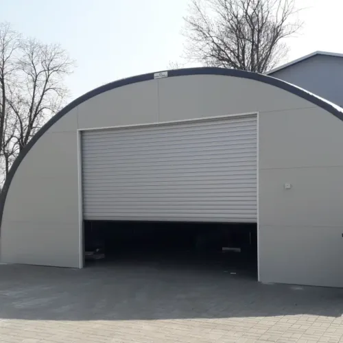 rzed halą z łukowym dachem widać częściowo otwartą bramę rolowaną prowadzącą do ciemnego wnętrza magazynu. Obiekt stoi na utwardzonym placu obok zabudowań gospodarczych.