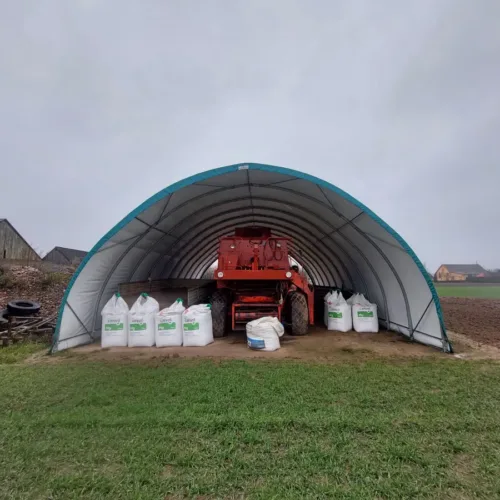 Hala namiotowa 10x15 ustawiona na skraju pola, wewnątrz rozsiewacz i big-bagi z nawozami. Lekka konstrukcja mobilna pozwala na szybkie zadaszenie sprzętu.