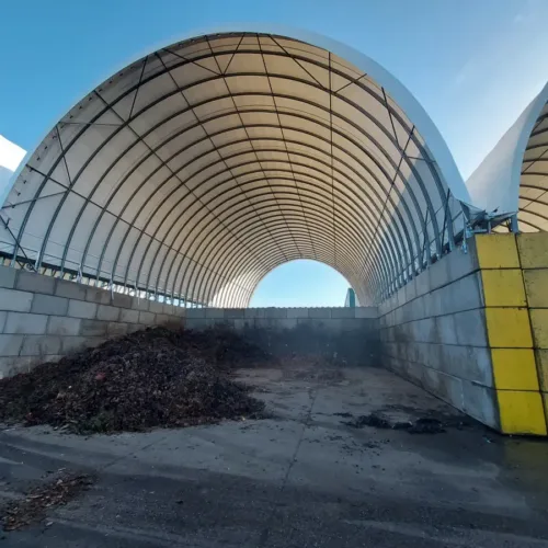 Wnętrze hali namiotowej o wymiarach 15x30 metrów z charakterystyczną łukową konstrukcją i jasnym dachem. Widoczne betonowe ściany z żółtymi elementami oraz materiał kompostowy składowany wewnątrz. Konstrukcja zapewnia dużą przestrzeń magazynową przy naturalnym doświetleniu.