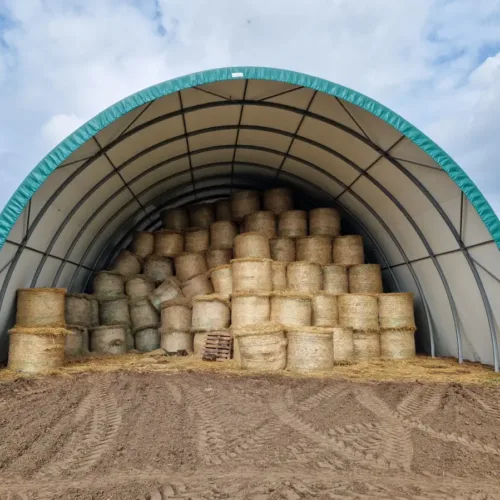 Hala namiotowa 15x30 metrów z turkusową membraną dachową, wypełniona stertami słomy ułożonymi pod łukową konstrukcją. Rolnicza hala magazynowa z solidną ramą stalową i pełnym zadaszeniem, chroniąca plony przed warunkami atmosferycznymi.