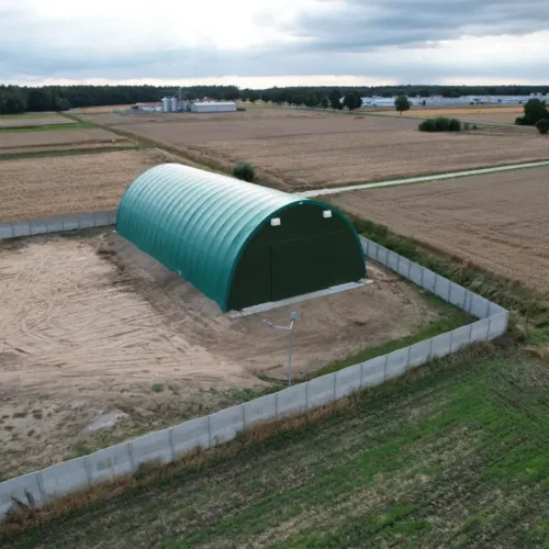 Ujęcie z drona przedstawiające halę namiotową 15x30 metrów w zielonym kolorze, położoną na terenie rolniczym otoczonym betonowym ogrodzeniem. Widoczna łukowa konstrukcja stalowa pokryta membraną PVC oraz otaczające pola uprawne i zabudowania gospodarcze w tle.