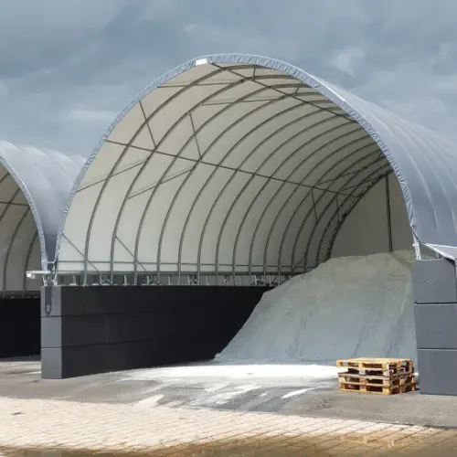 Dwie hale namiotowe 10x15 na placu komunalnym, jedna wypełniona pryzmą soli drogowej. Ściany oporowe z bloków i wysoki prześwit ułatwiają załadunek.