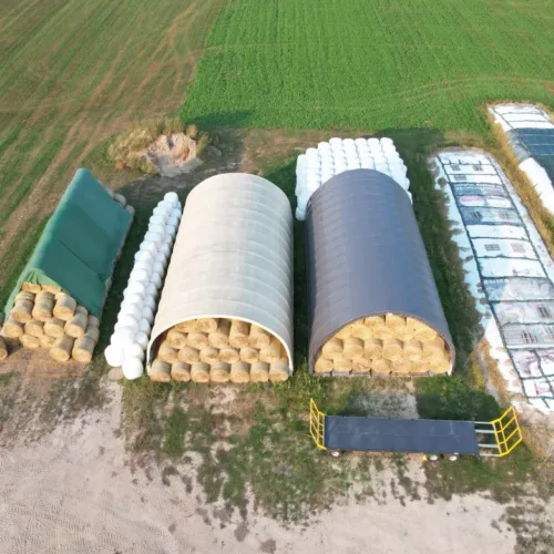 Dwie hale namiotowe 10x20 w gospodarstwie, wypełnione belami słomy ułożonymi w piramidy. Z boku widać rzędy sianokiszonek i plac manewrowy.