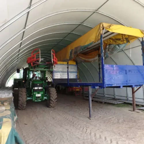 Namiot garażowy 10x36x5x1,5 m z ciągnikiem i przyczepami. Długa hala namiotowa z betonową posadzką do składowania sprzętu.