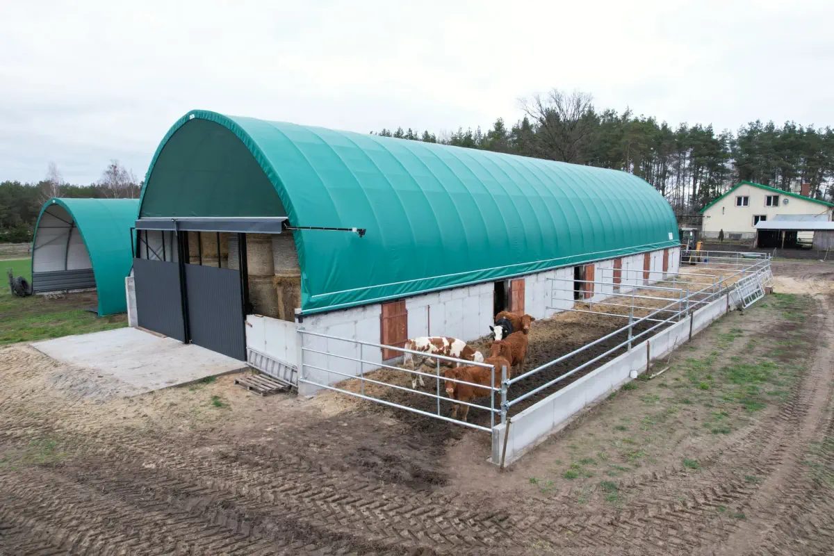 Hala namiotowa posadowiona na bloczkach betonowych, z zielonym poszyciem w kształcie łuku.