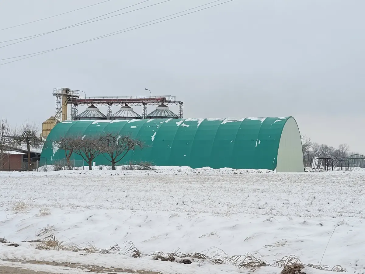 Duża hala z zielonym poszyciem membranowym na zaśnieżonym terenie w sąsiedztwie silosów zbożowych. Zadaszenie zapewnia ochronę materiałów i pracę w trudnych warunkach zimowych.